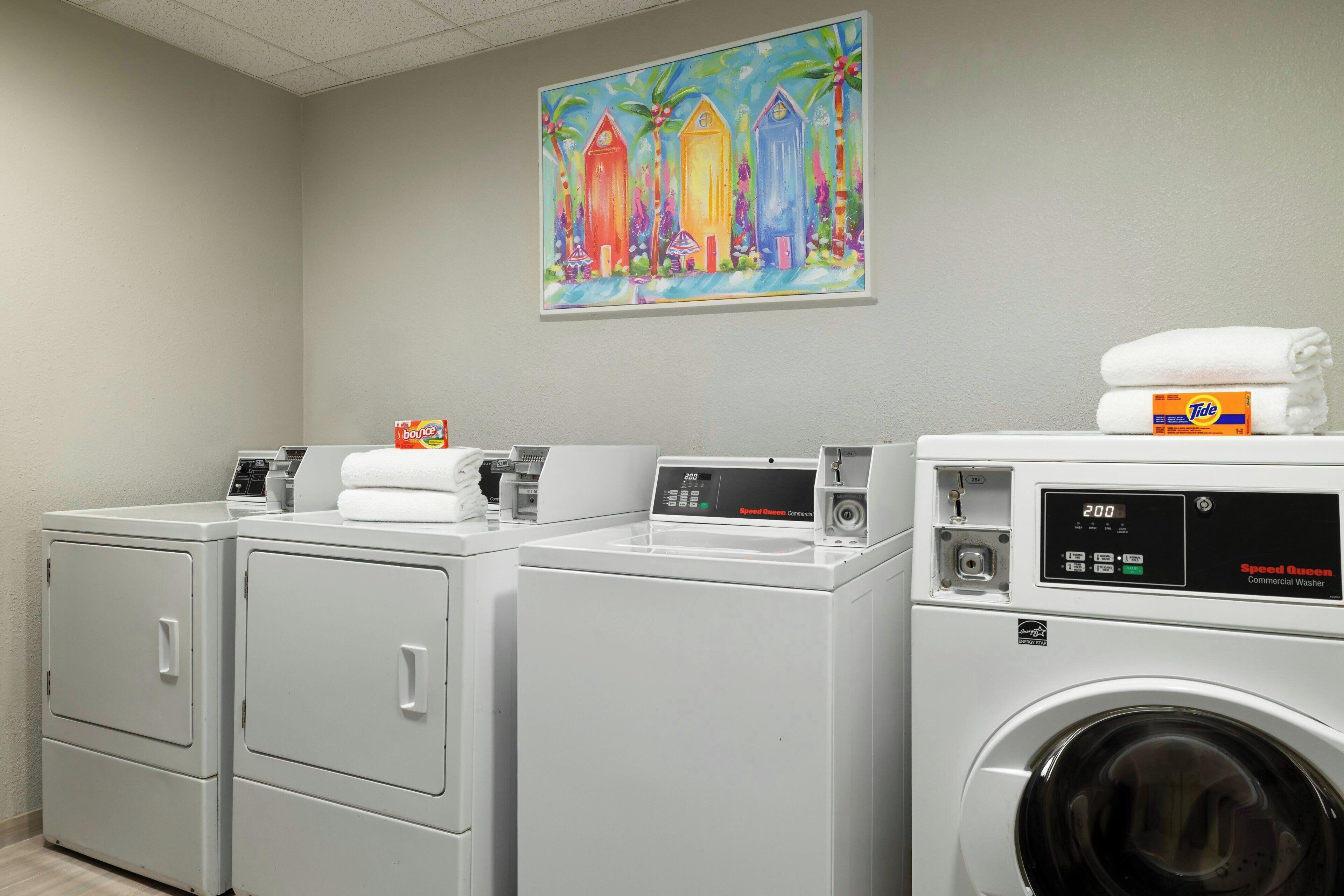A laundry area with three washers and dryers, a painting of colorful buildings, and a Tide detergent box on the dryer.