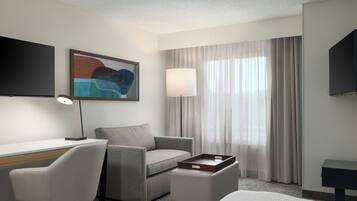 Habitación de hotel moderna con televisor de pantalla plana, escritorio, sillón cómodo, mesita de noche y un amplio ventanal con cortinas.