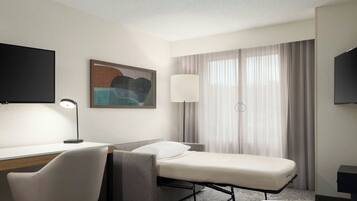 A hotel room with a desk, chair, bed, TV, lamp, and curtains.
