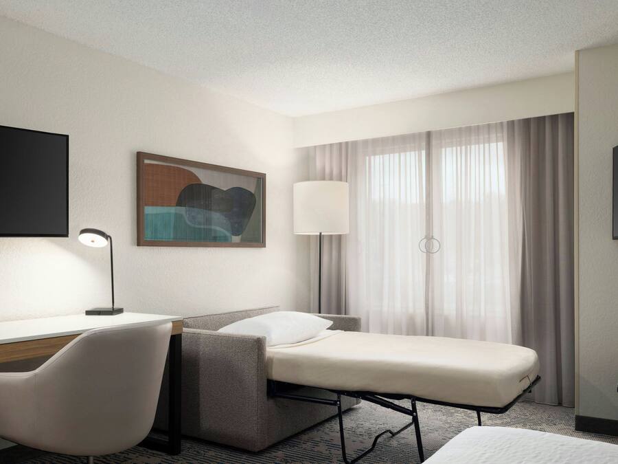 A hotel room with a desk, chair, bed, TV, lamp, and curtains.