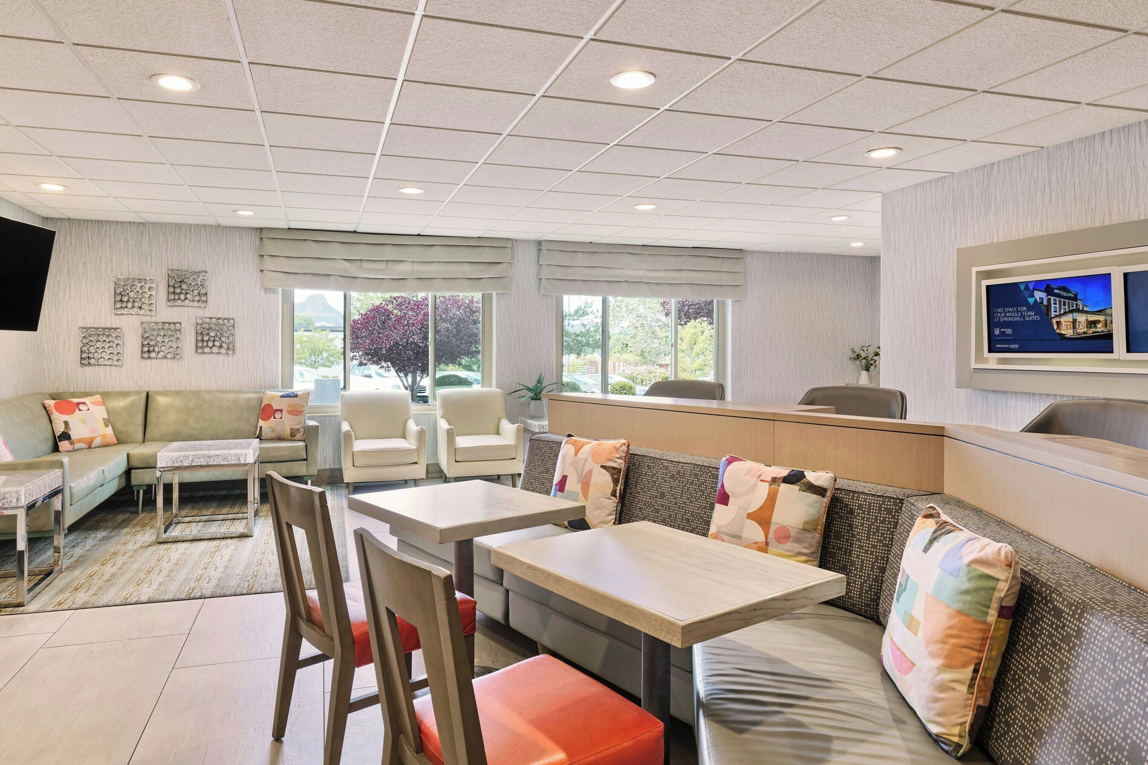 A modern hotel room with a sofa, two tables, a TV, and a window with a view of trees.