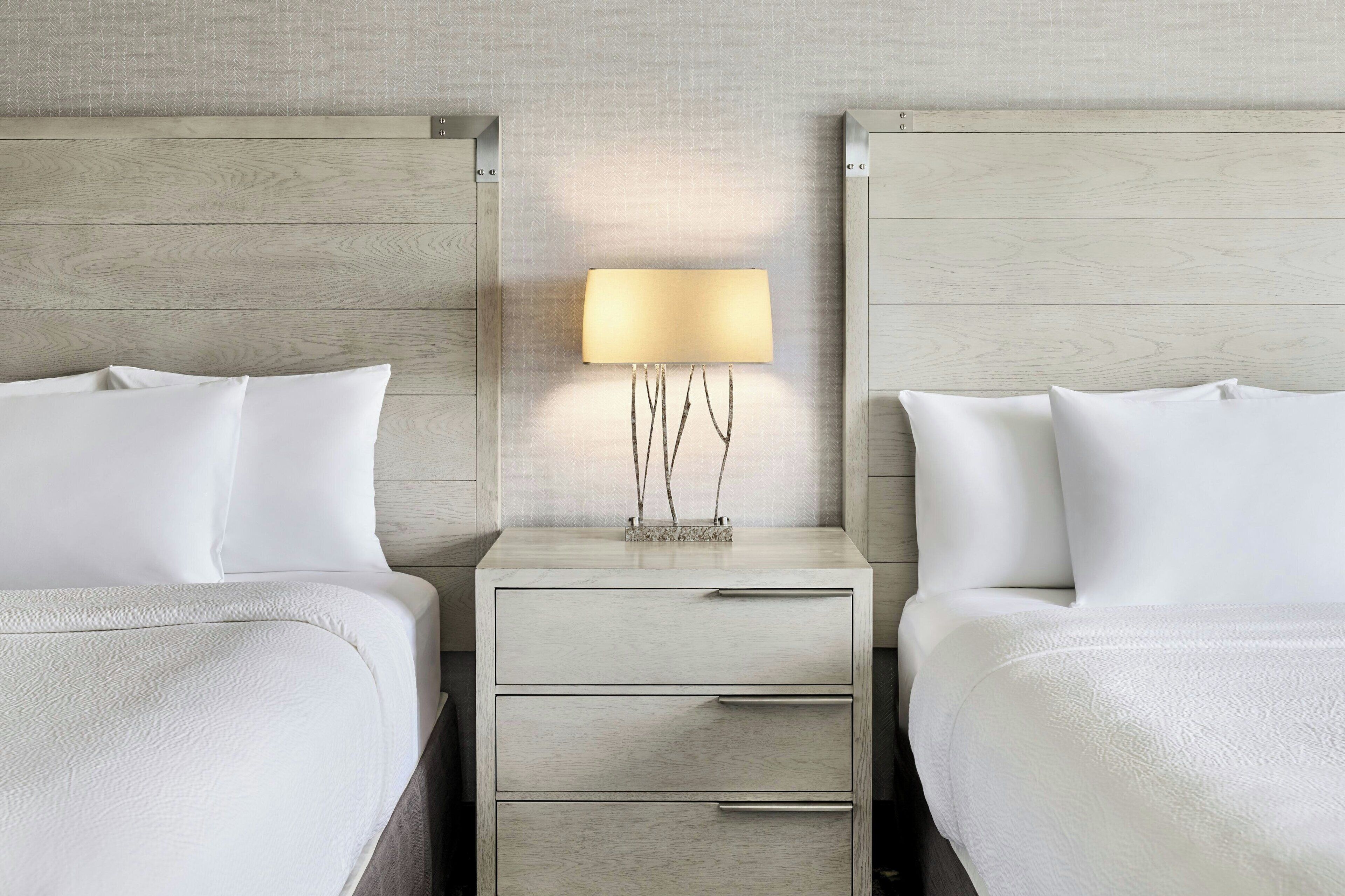 A hotel room with two beds, a nightstand with a lamp, and a small table with a vase.