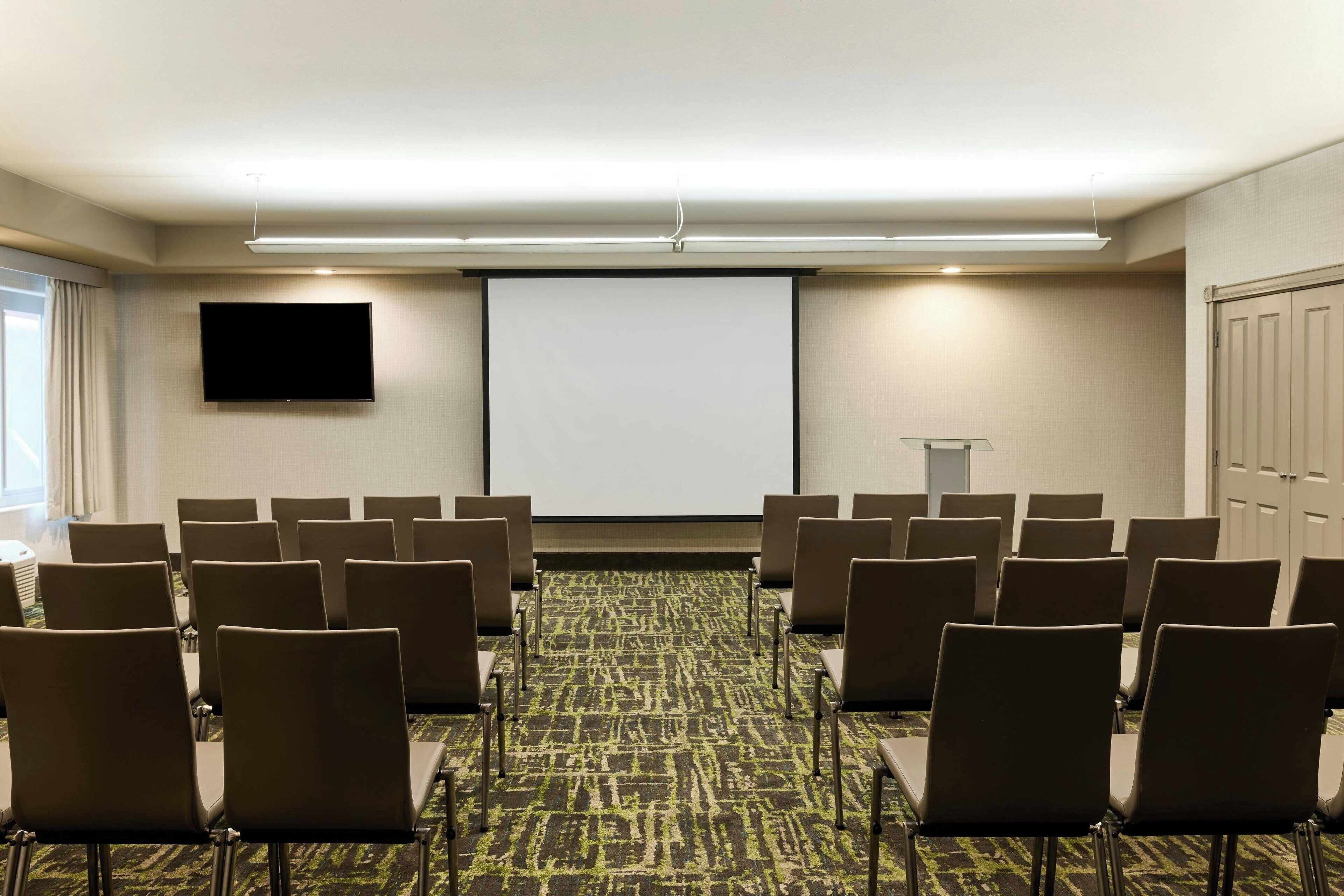 A conference room with rows of chairs, a projector screen, and a flat-screen TV on the wall.