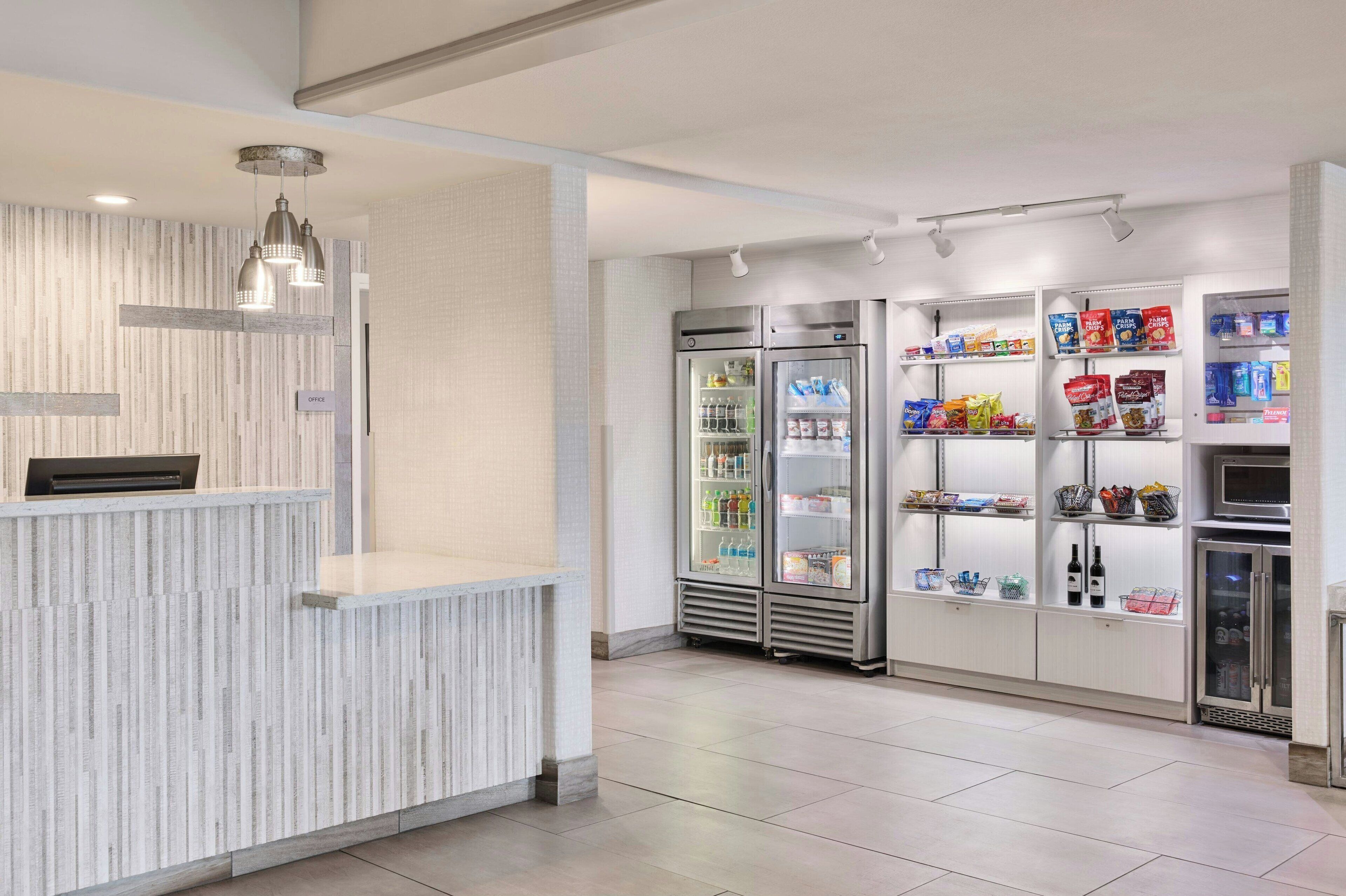 A clean, modern reception area with a reception desk, two refrigerators stocked with beverages, shelves with snacks, and a microwave.