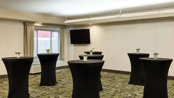 Uma sala de conferência com quatro mesas redondas cobertas por toalhas pretas, cada uma com uma pequena planta em cima. Há uma televisão de tela plana na parede e uma janela com cortinas à esquerda.