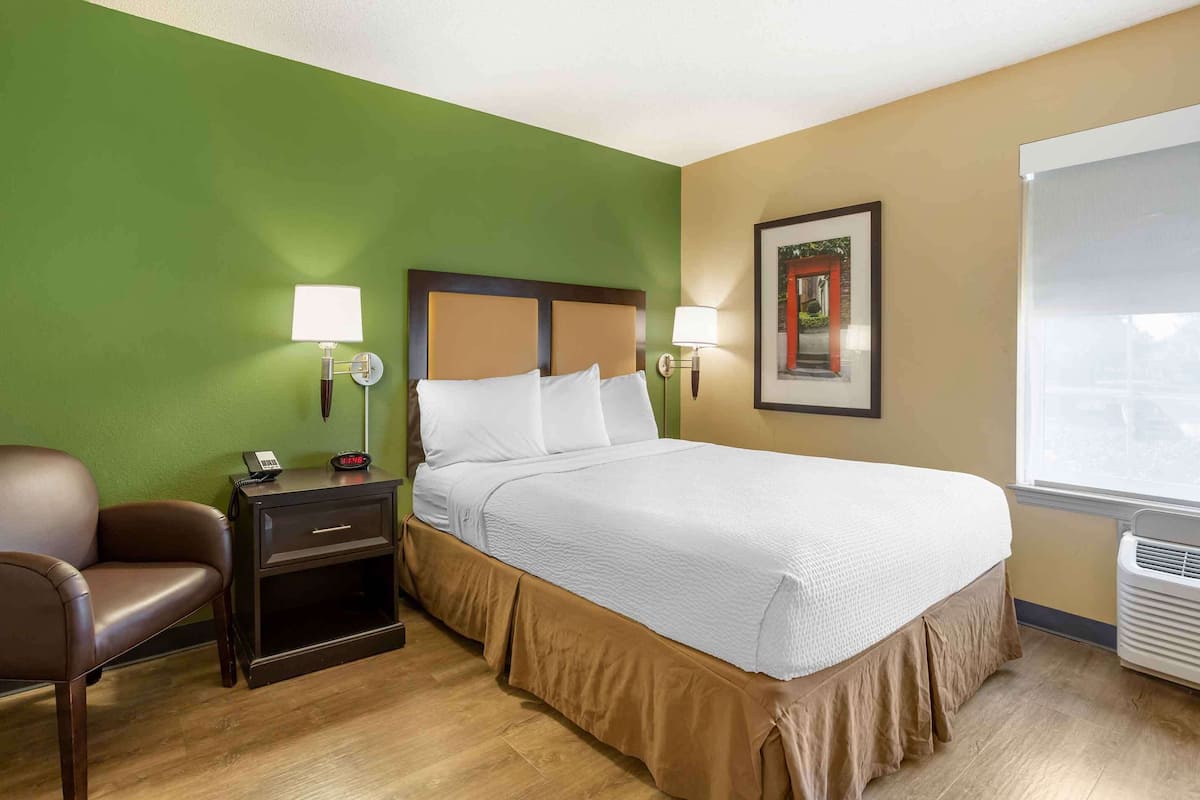 A hotel room with a bed, bedside lamps, a chair, a nightstand, a framed picture on the wall, and a window.