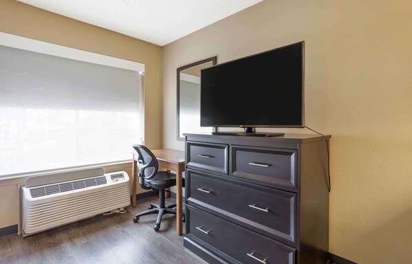A room with a large window, a desk, a chair, a dresser, and a flat-screen TV.