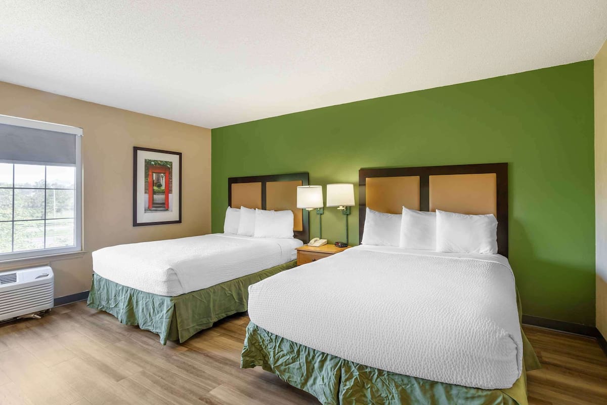 A hotel room with two beds, green walls, a framed picture, bedside lamps, and a window with a view.