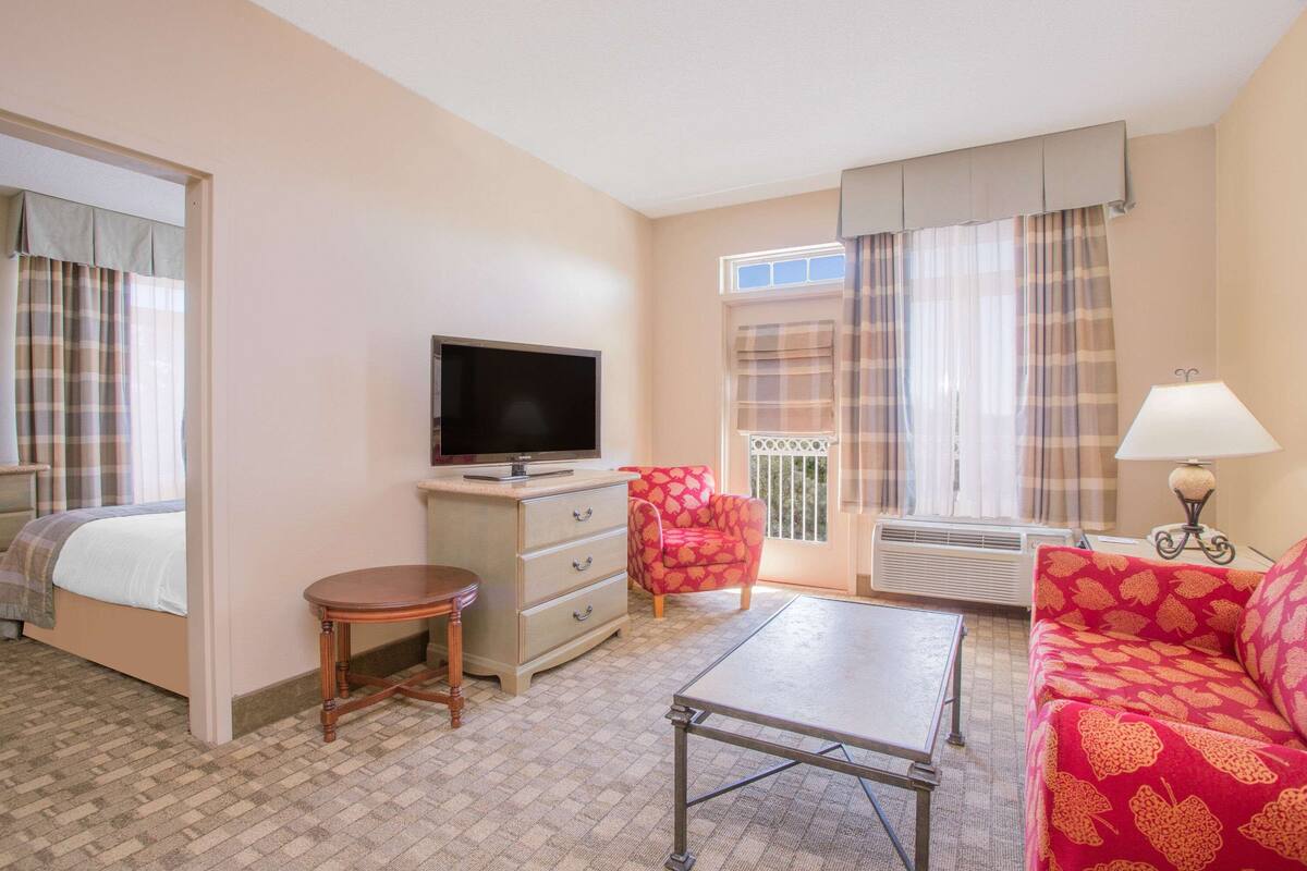 Quarto de hotel com cama, televisão, escrivaninha, poltrona, sofá e janela com cortinas.