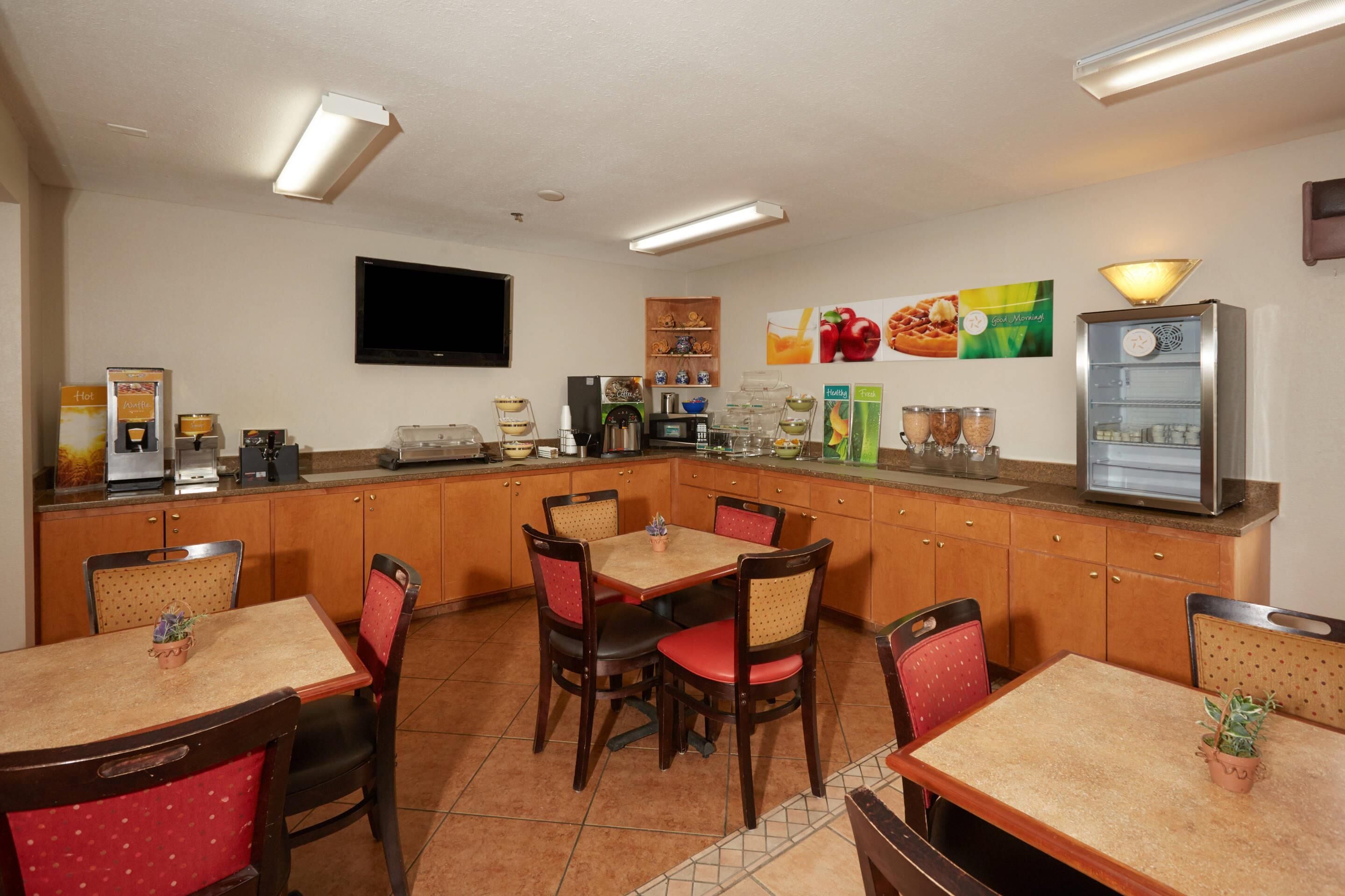 A hotel dining area with wooden tables and chairs, a kitchen counter with appliances, and a refrigerator.