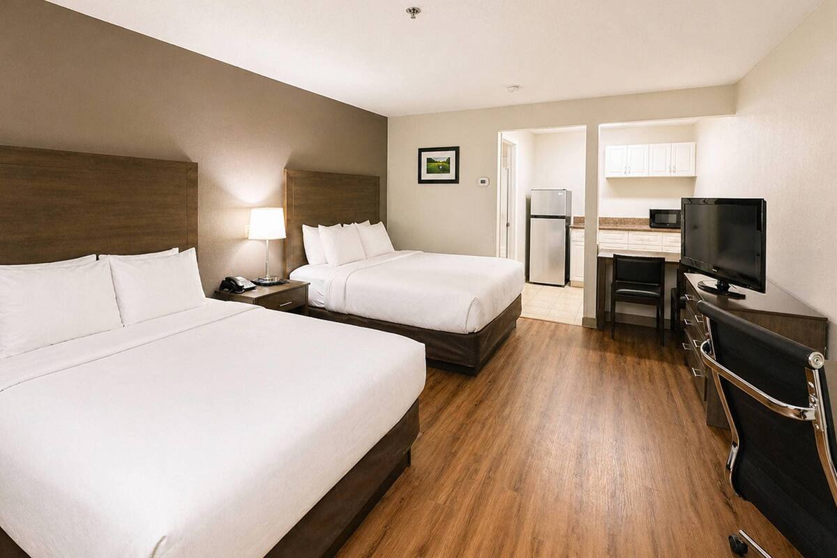 A hotel room with two beds, a desk with a chair, a television, and a kitchenette.