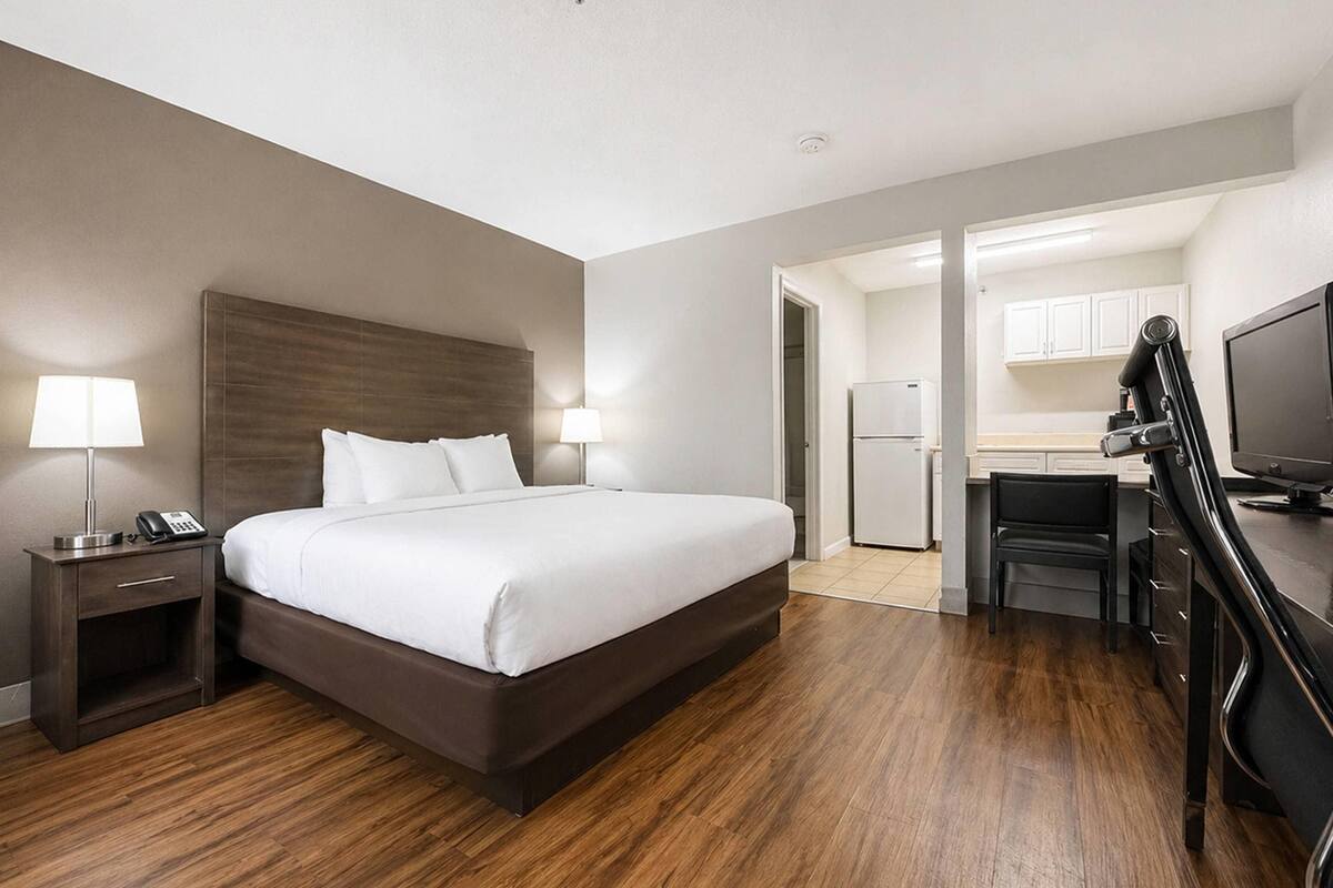 A hotel room with a bed, bedside tables, a desk, a chair, a television, and a kitchenette.