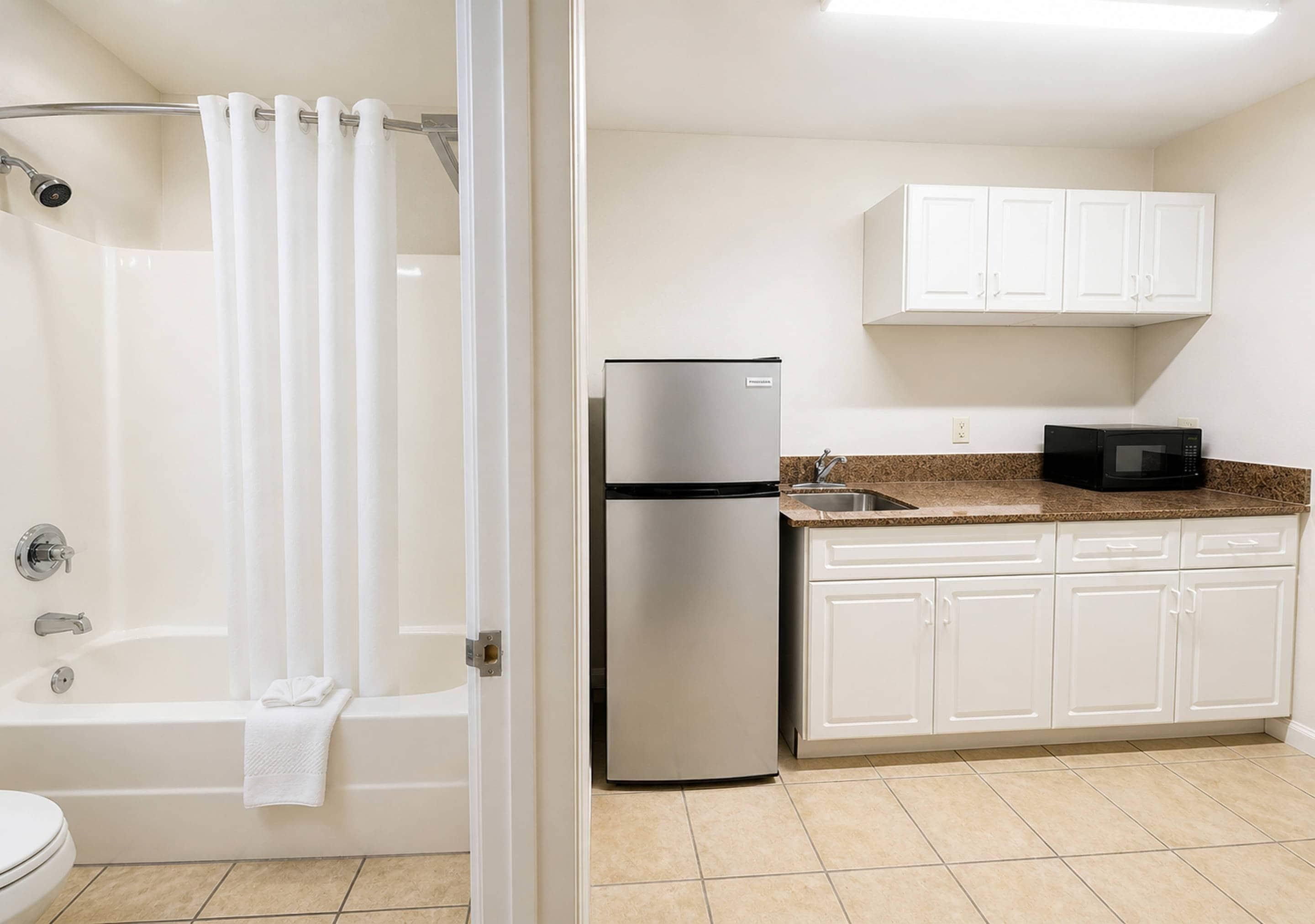 A bathroom with a bathtub, shower, and toilet. Adjacent is a kitchen with a refrigerator, microwave, and white cabinets.