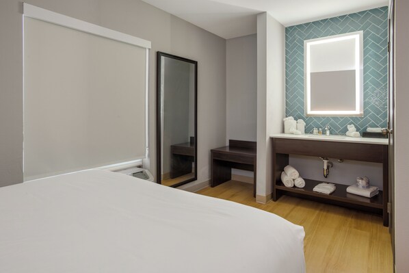 A hotel room with a bed, a mirror, a vanity with a sink, and a shelf with towels.