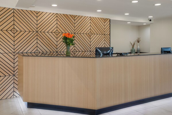 Una recepción con un muro de diseño geométrico, un jarrón con flores naranjas y un monitor de computadora.