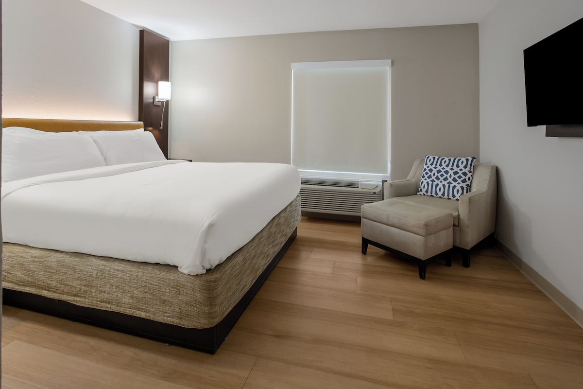 A hotel room with a large bed, a small sofa, a flat-screen TV, and a window with blinds.