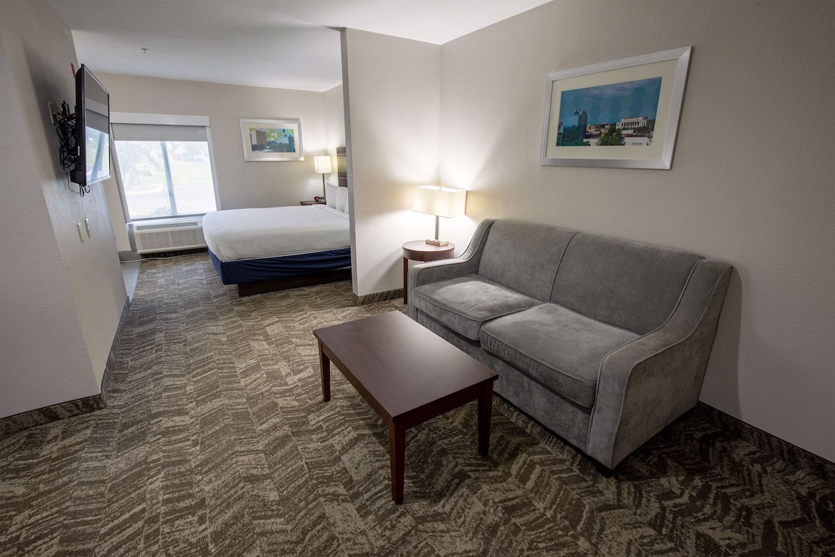 A hotel room with a bed, a grey sofa, a wooden coffee table, and a lamp.