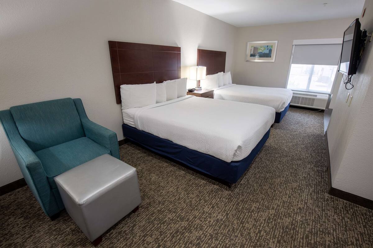 A hotel room with two beds, a teal armchair, a white ottoman, a nightstand with a lamp, and a TV mounted on the wall.