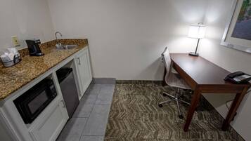 Cocina compacta con microondas, cafetera y lavabo. Hay un escritorio pequeño con lámpara y teléfono. El piso está alfombrado.