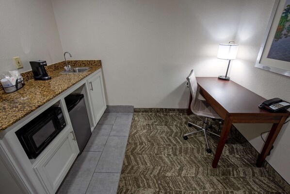 Cocina compacta con microondas, cafetera y lavabo. Hay un escritorio pequeño con lámpara y teléfono. El piso está alfombrado.