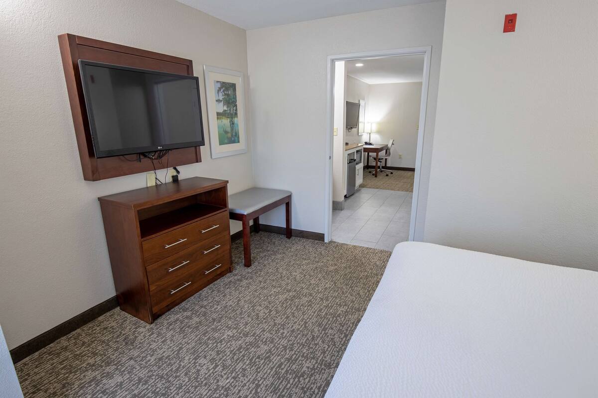 A hotel room with a large flat-screen TV mounted on the wall, a wooden dresser with drawers, a small bench, and a bed with a white comforter.
