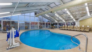Una piscina interior con paredes de vidrio transparente, una escalera de piscina y varias sillas alrededor del área de la piscina.