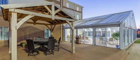 An outdoor patio with a wooden pergola, a glass greenhouse, and a pool area.