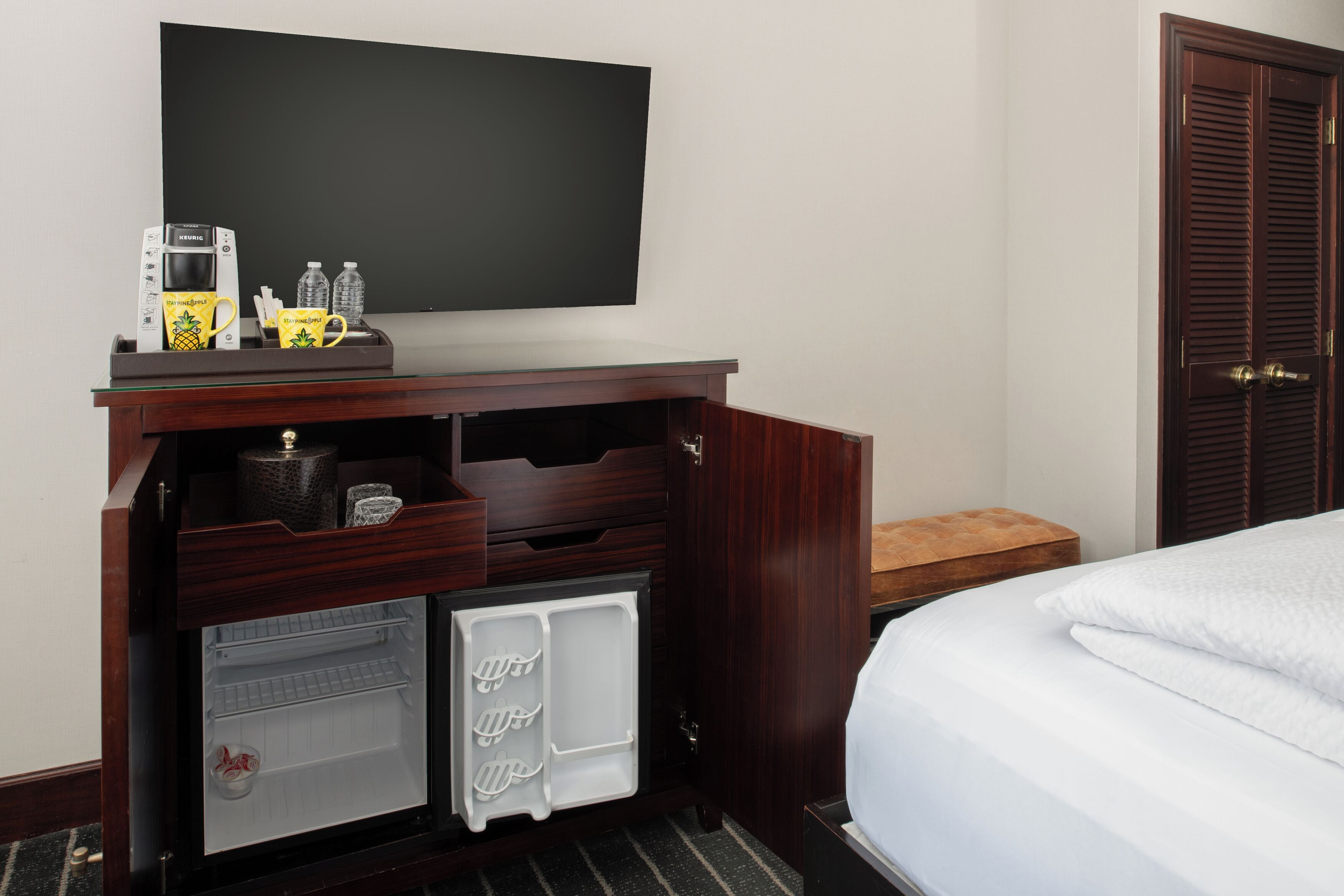 Una habitación de hotel con una cama, televisión montada en la pared, un minibar y una puerta de madera.