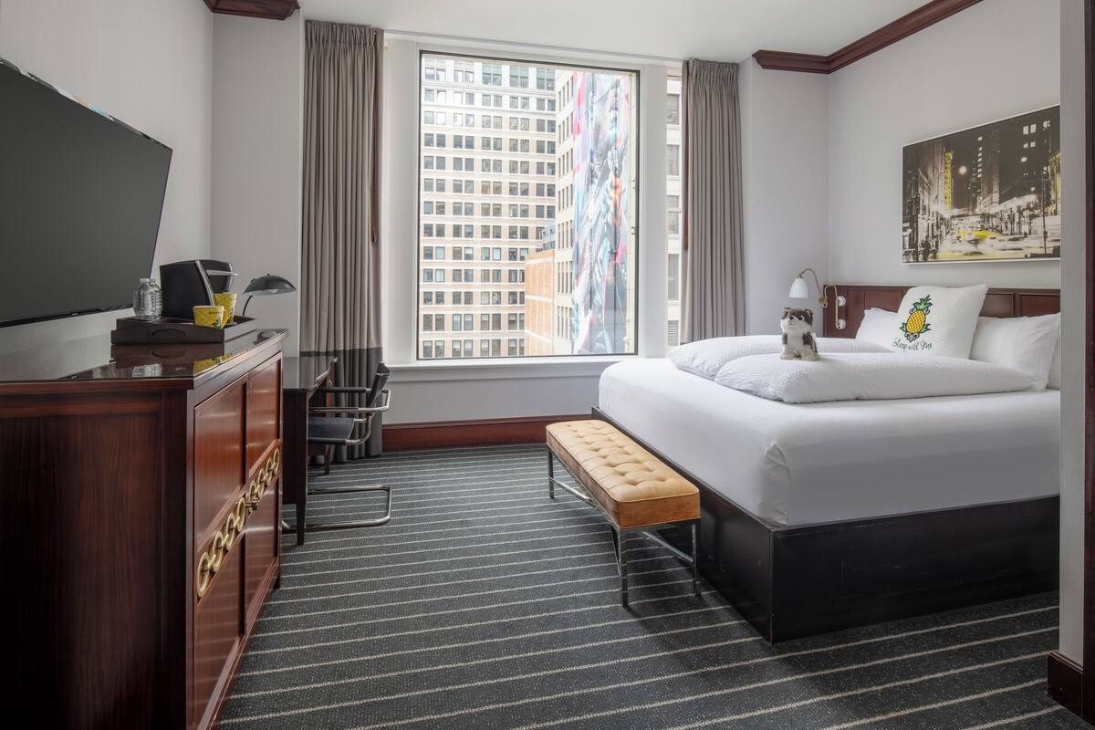 A hotel room with a large bed, a desk with a flat-screen TV, a bench, and a window with a city view.