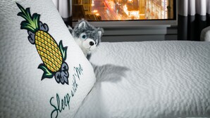 Una cama con un almohadón que presenta un diseño de piña y el texto "Sleep with me". Una ventana muestra un paisaje urbano nocturno.