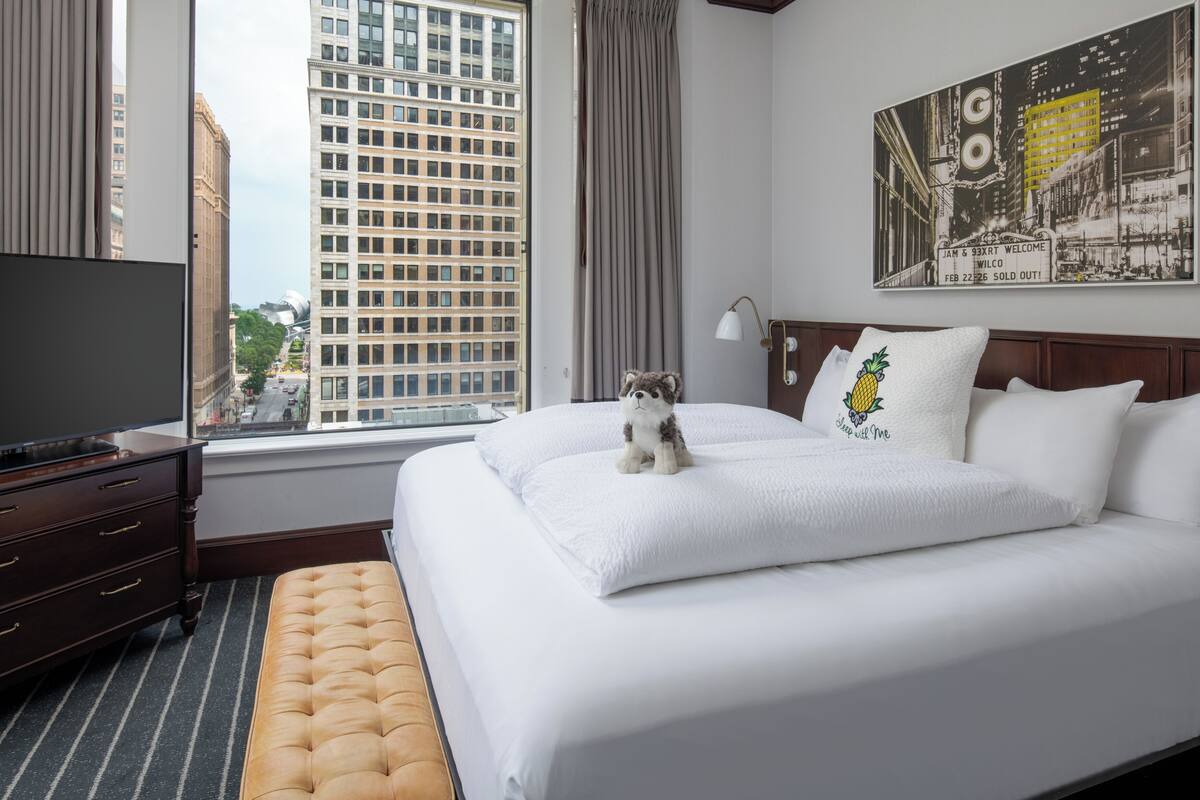 A hotel room with a large bed, a television, a window with curtains, a city view, and a painting on the wall.