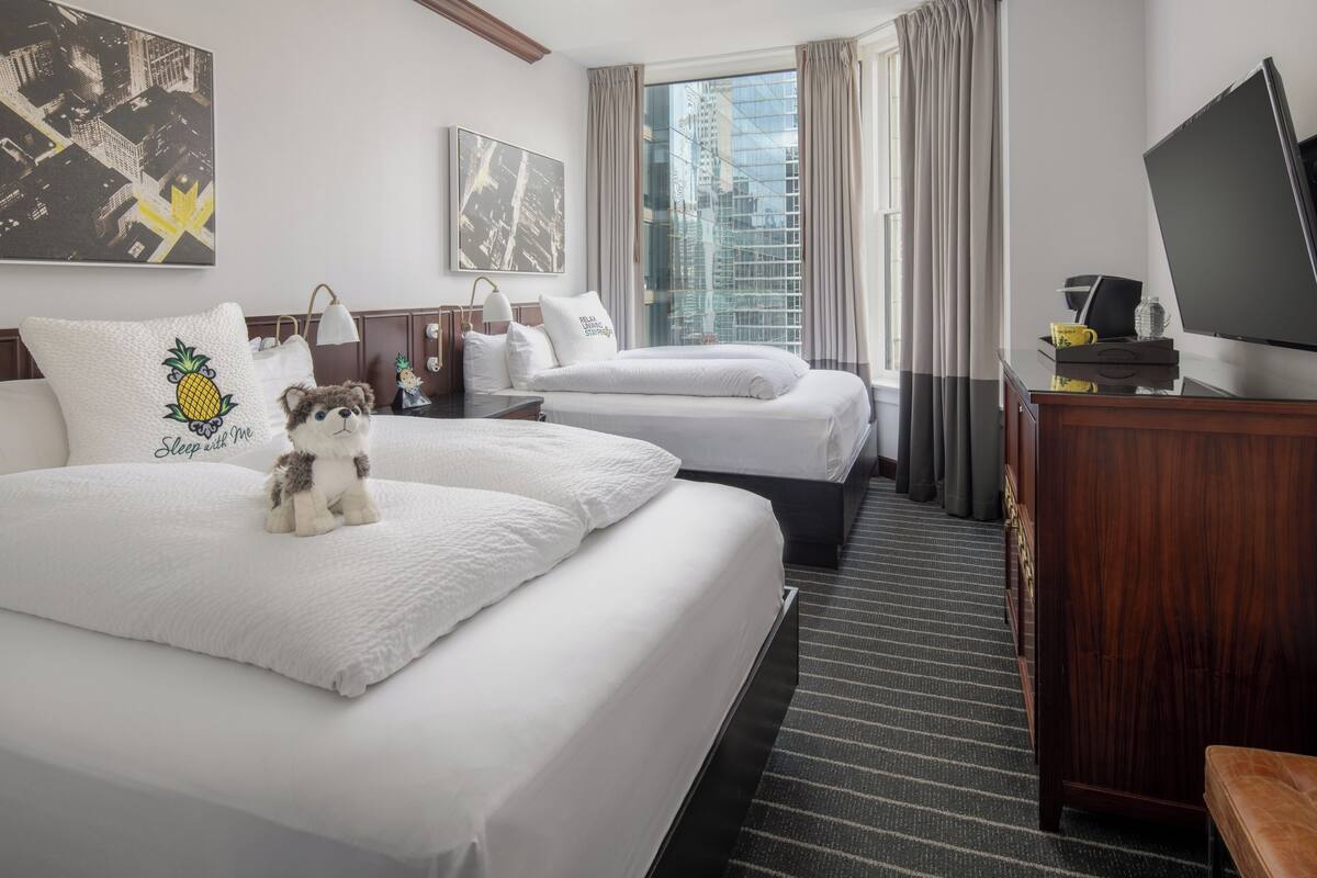 A hotel room with two beds, a TV, a desk, and a window with a city view.
