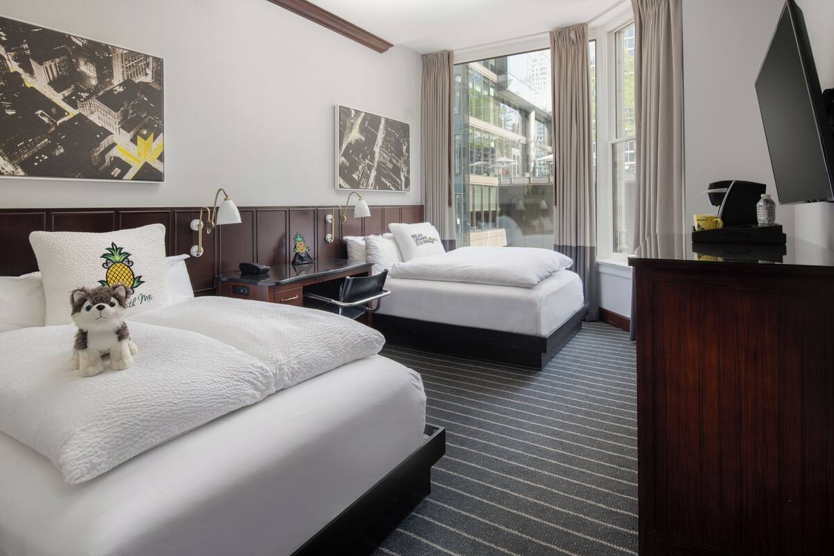 A hotel room with two beds, a desk, a TV, and large windows with curtains.