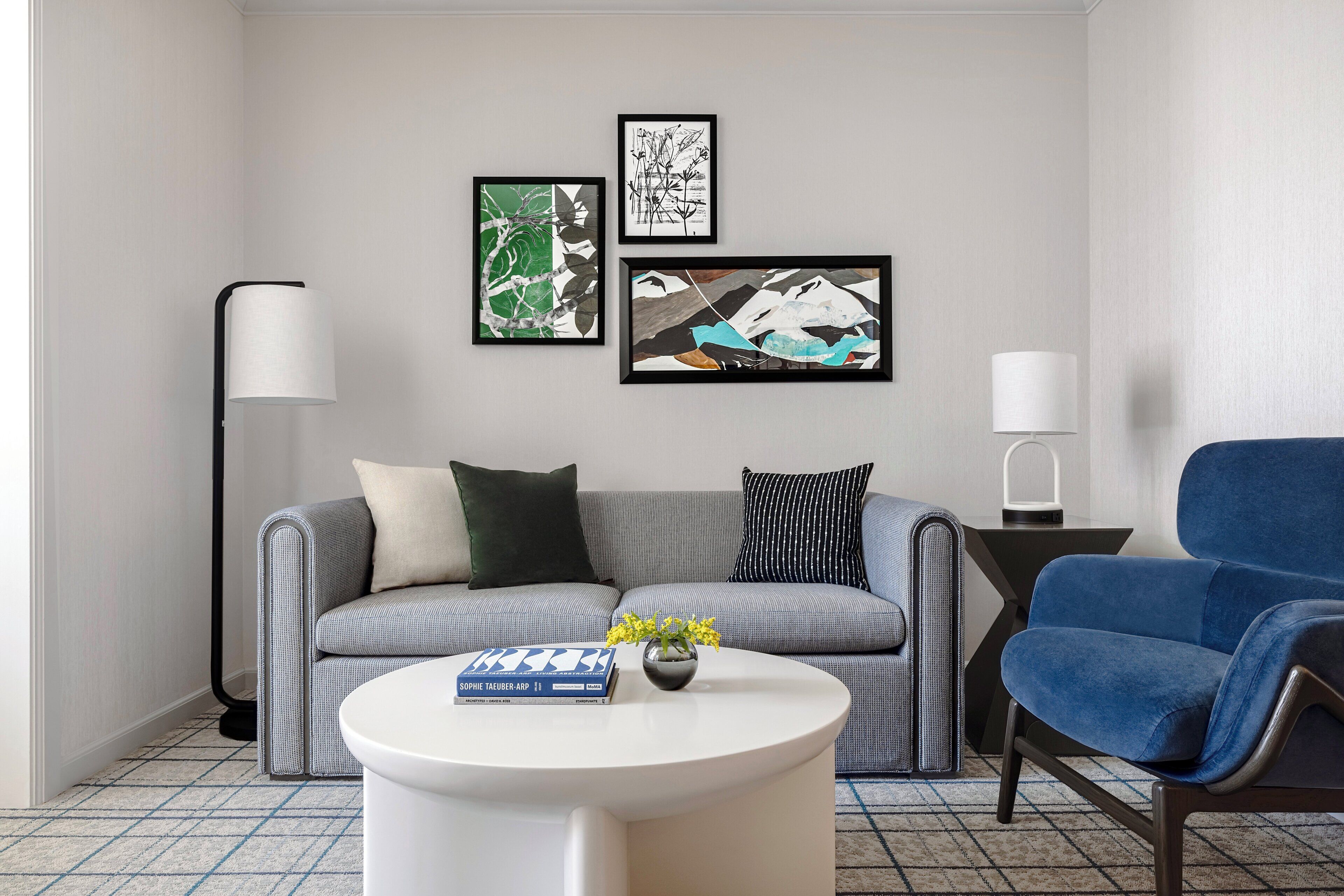 Una sala de estar moderna con un sofá gris, un sillón azul, una mesa de centro blanca y tres obras de arte enmarcadas en la pared.