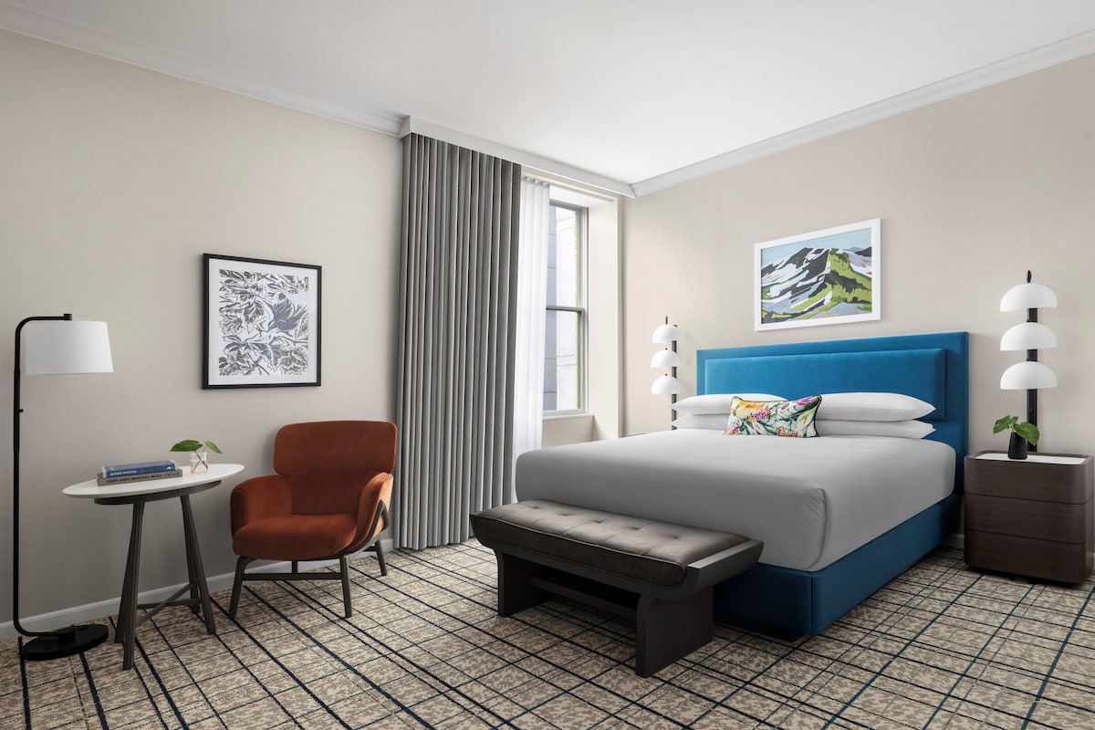 A hotel room with a bed, a chair, a small table, a lamp, and a picture on the wall.