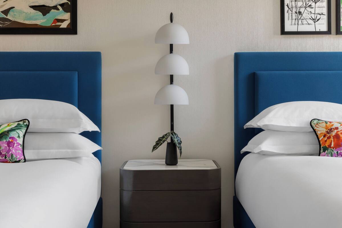 Ein Hotelzimmer mit zwei Betten, beide mit blauen Kopfteilen, weißer Bettwäsche und blumenmusterten Kissen. Ein schwarzer Nachttisch mit Blumenvase steht zwischen den Betten.