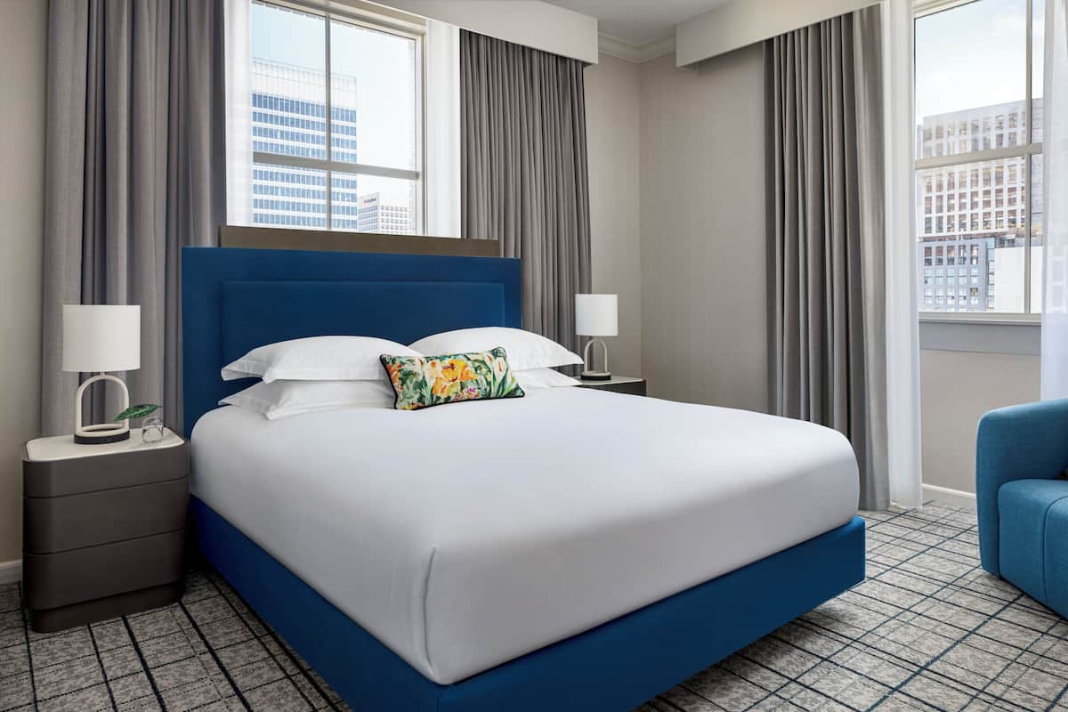 Ein Hotelzimmer mit einem großen Bett, zwei Nachttischen, einem Beistelltisch, einem blauen Sessel und einem Fenster mit Vorhängen.