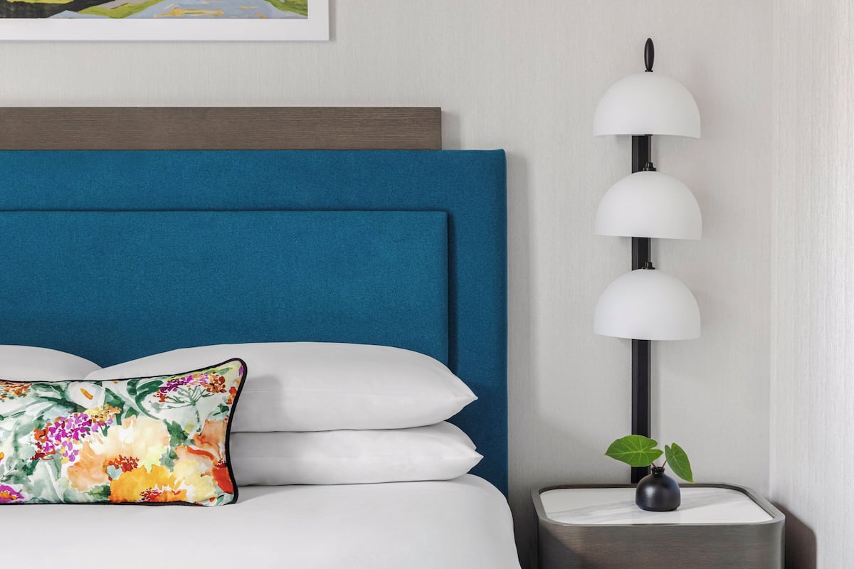 Ein ordentlich bezogenes Bett mit blauem Kopfteil, weißen Kissen und einem farbenfrohen Blumenkissen. Neben dem Bett steht eine hohe, moderne Lampe mit mehreren Schirm und auf dem Nachttisch ein kleiner Blumenstrauß in einer Vase.