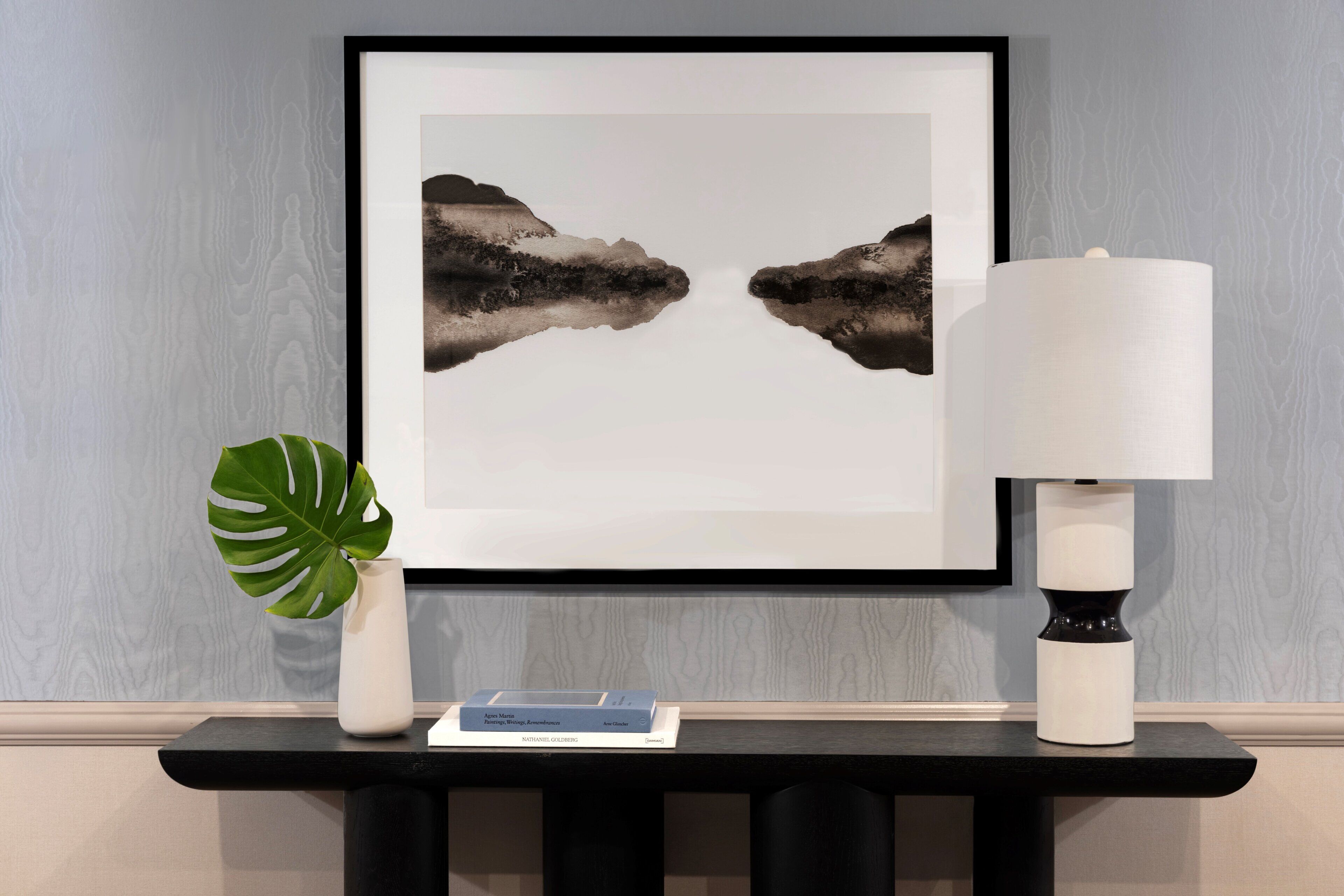 A framed artwork on a wall, a white vase with a plant, and a table with a lamp and a book.