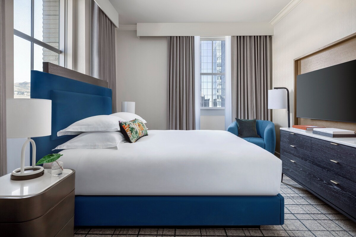Habitación de hotel moderna con una cama grande, cabecera azul, televisor de pantalla plana, cómoda y ventana con cortinas.