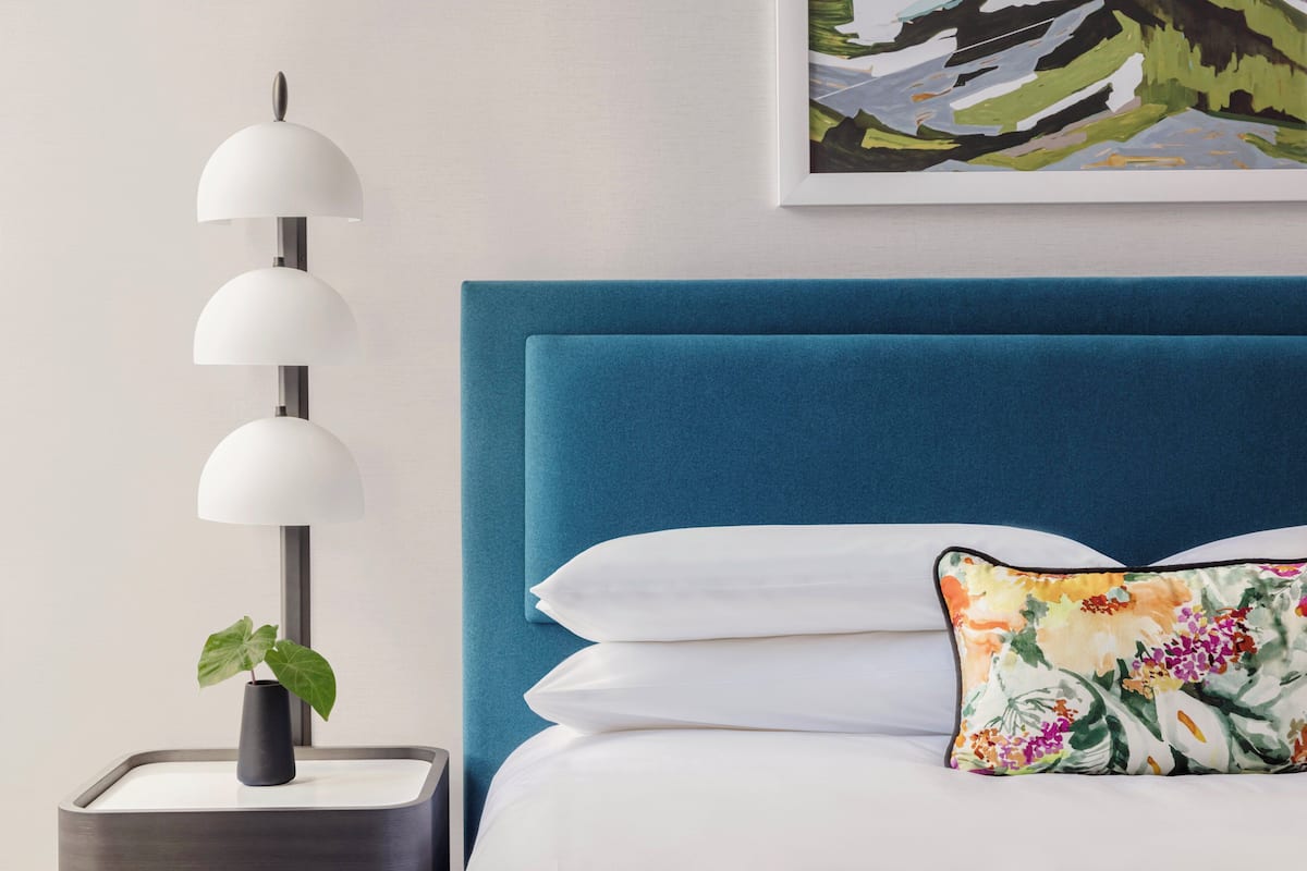 Ein ordentlich eingerichtetes Schlafzimmer mit blauem Kopfteil, einer Nachttischlampe und einem blumigen Kissen.