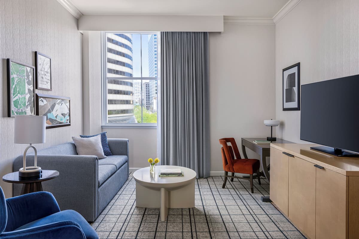 Ein modernes Hotelzimmer mit grauem Sofa, einem runden Couchtisch, einem Fernseher und Blick auf Gebäude durch ein großes Fenster.
