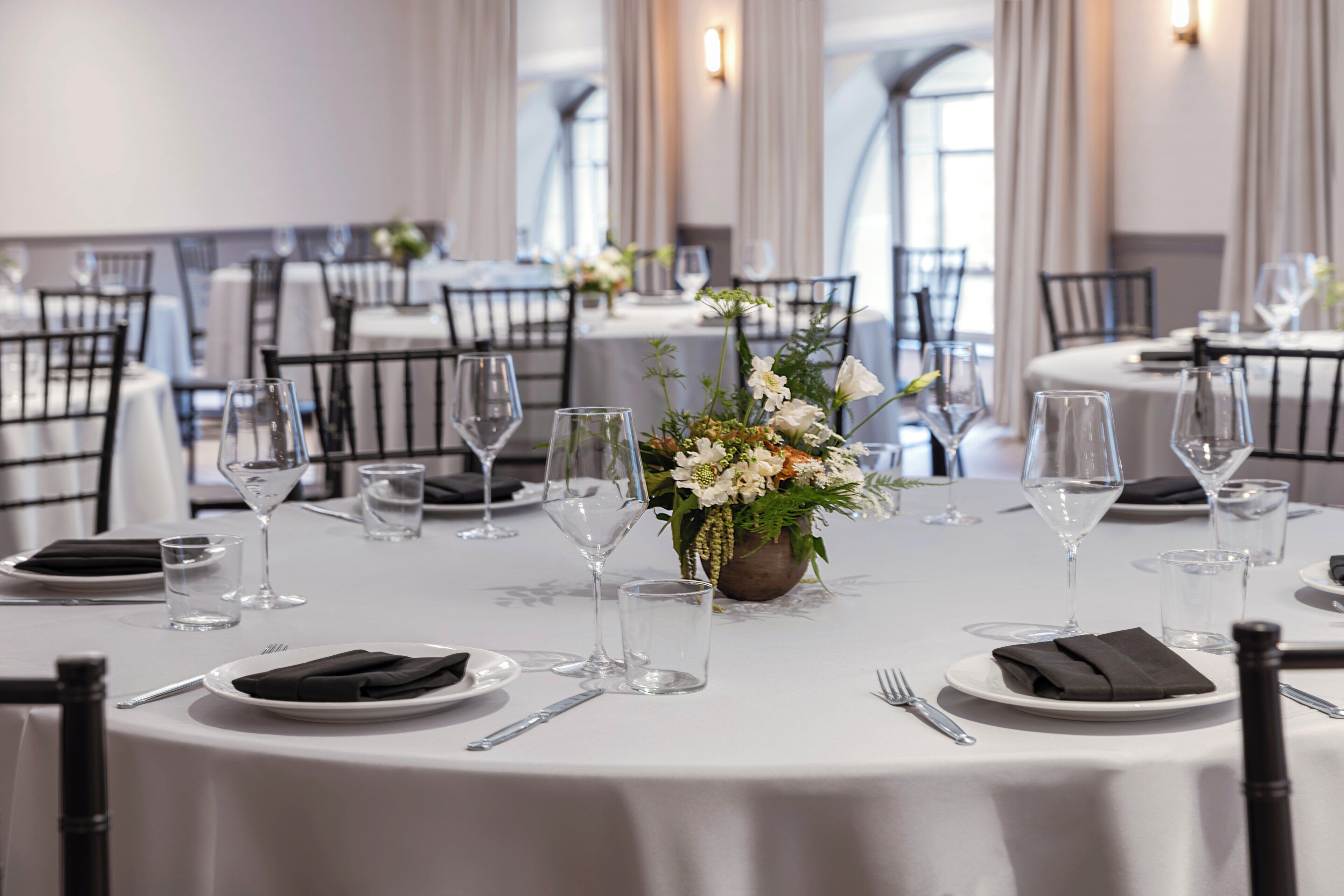A dining area with round tables set for a meal, each with a white tablecloth, black chairs, and a centerpiece of white flowers.