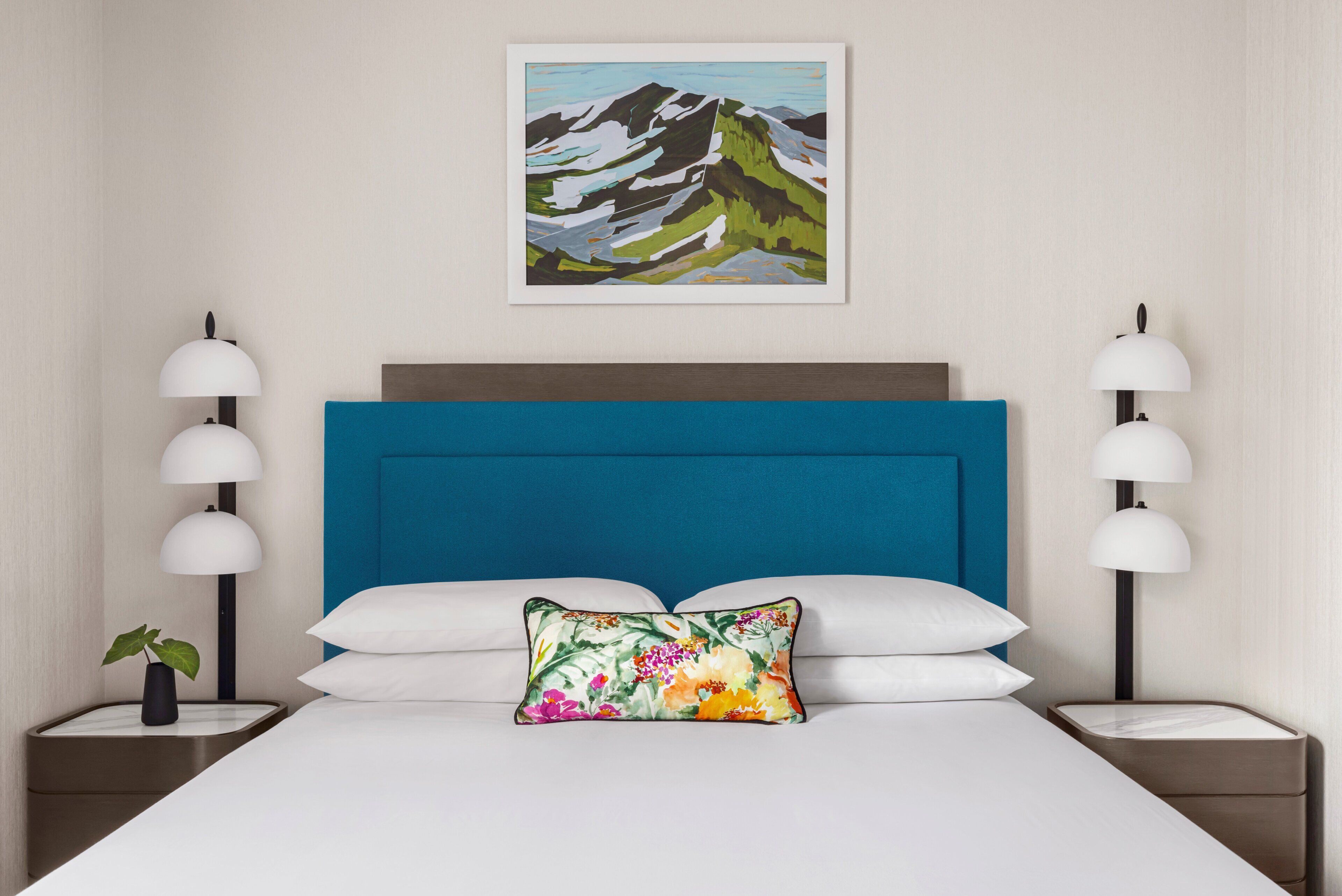 A hotel room with a large bed, a blue headboard, white bedding, and a floral pillow. There are two bedside tables with lamps and a small plant on one.