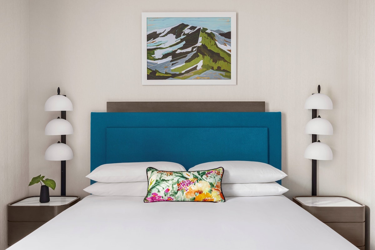 A hotel room with a large bed, a blue headboard, white bedding, and a floral pillow. There are two bedside tables with lamps and a small plant on one.