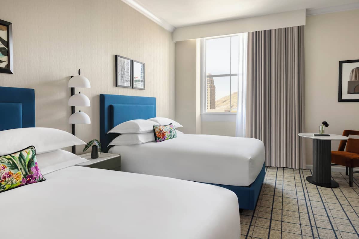 Ein Hotelzimmer mit zwei Betten, einem kleinen Tisch, einem Stuhl, einem Fenster mit Vorhängen und Blick auf ein Gebäude.