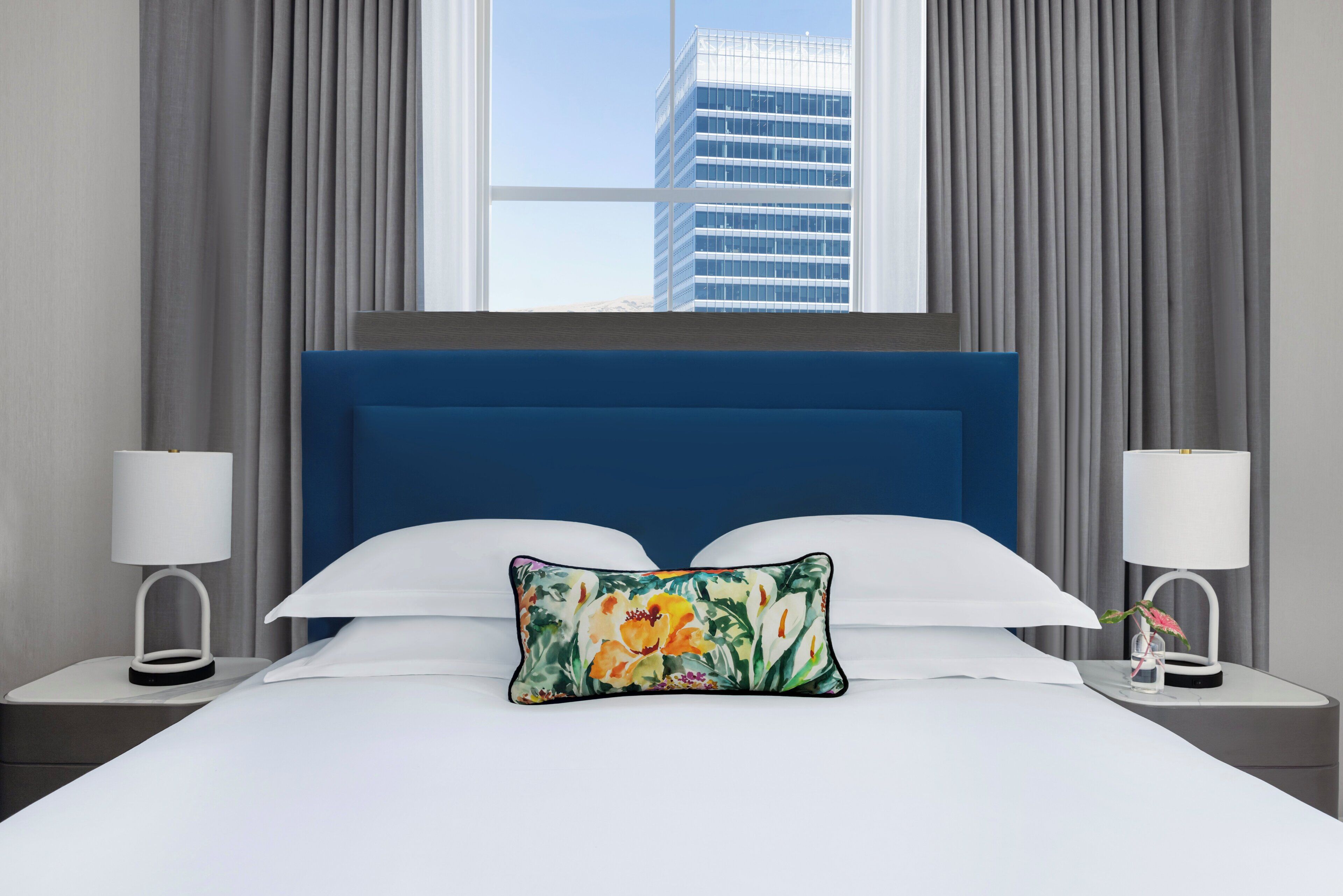 A hotel room with a large bed, a blue headboard, white bedding, and a floral pillow. There are two bedside tables with lamps and a window with curtains.