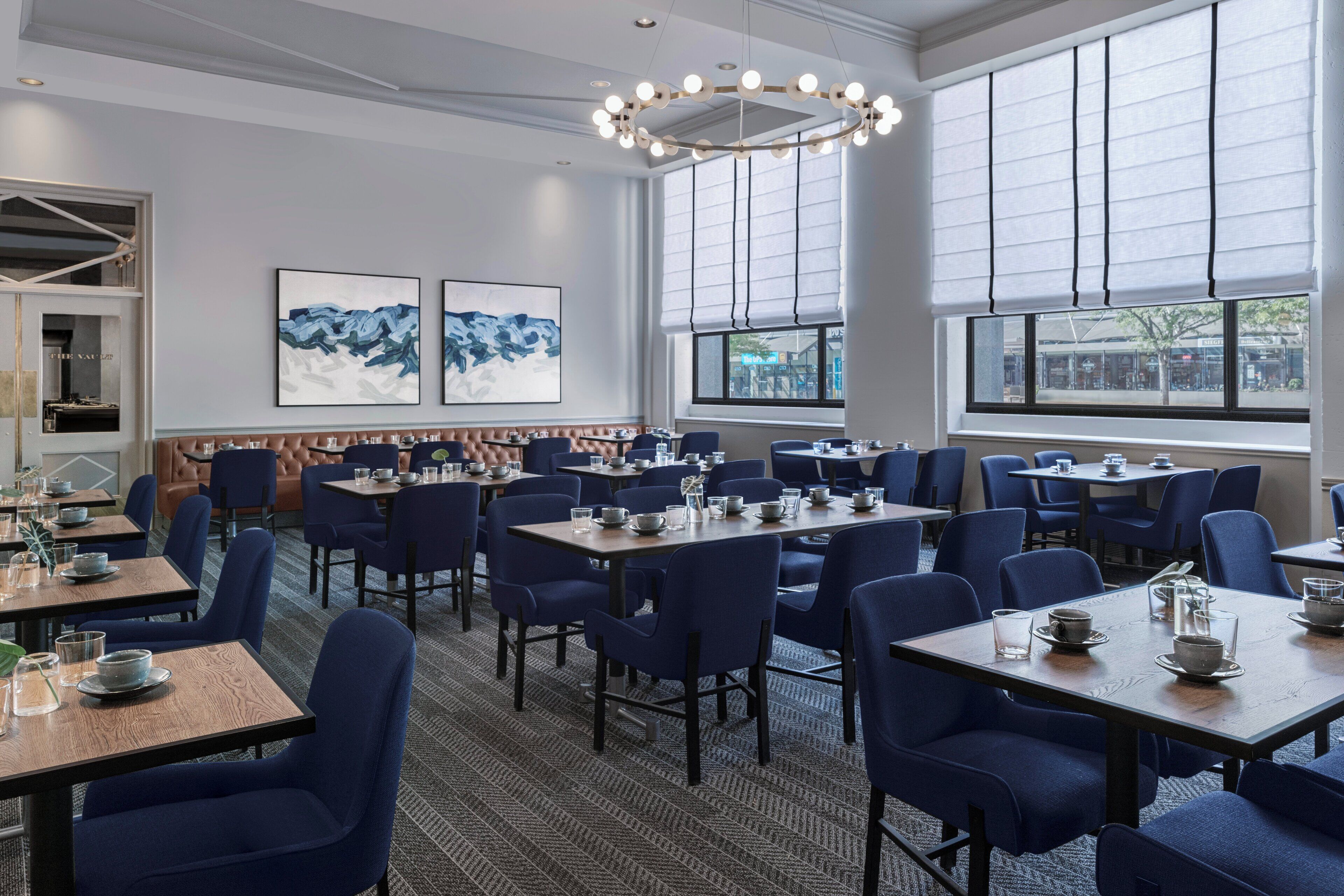 A hotel dining area with tables and chairs, artwork on the walls, and large windows.