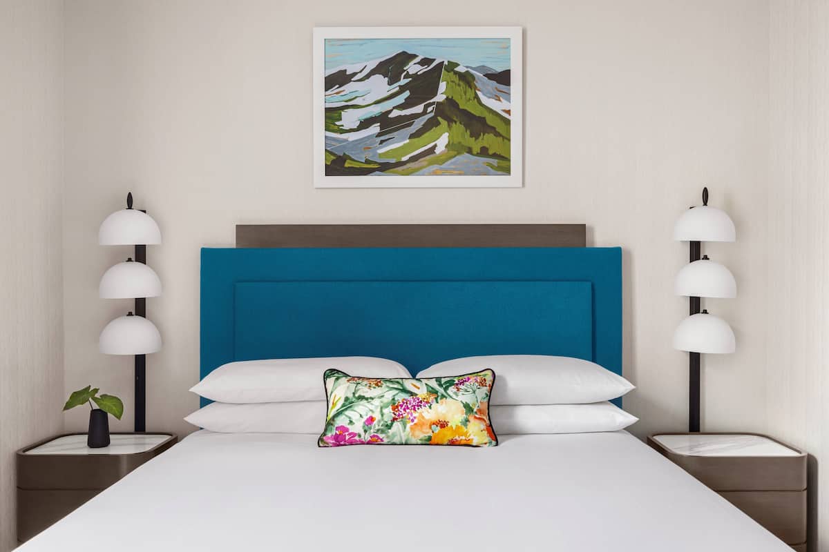 Ein Hotelzimmer mit einem großen Bett, einem blauen Kopfteil, weißer Bettwäsche und einem Blumenkissen. Es gibt zwei Nachttische mit Lampen und eine kleine Pflanze auf einem.
