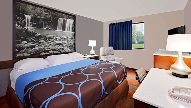 Una habitación de hotel con una cama grande, un escritorio con teléfono, una silla, una mesita de noche con lámpara y un ventanal con cortinas.